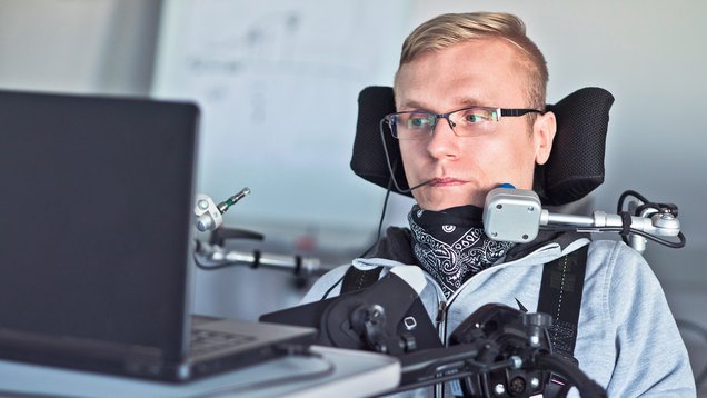 Foto eines Mannes mit blonden Haaren, der im Rollstuhl sitzt und mittels Mundstab und Trackball Maus versucht einen Computer zu bedienen.
